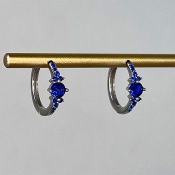 To sølvfarvede creoler med blå zirkonia hænger på en gylden stang.